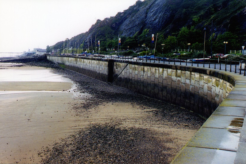 A construction view of the A6 Manchester Airport Relief Road project, showcasing a large retaining wall built using Geoquest Co. Ltd.'s precast concrete facing panels. The image highlights the integration of the road with surrounding infrastructure, illustrating the scale of the project and the innovative design solutions employed for the retaining walls and bridge abutments.