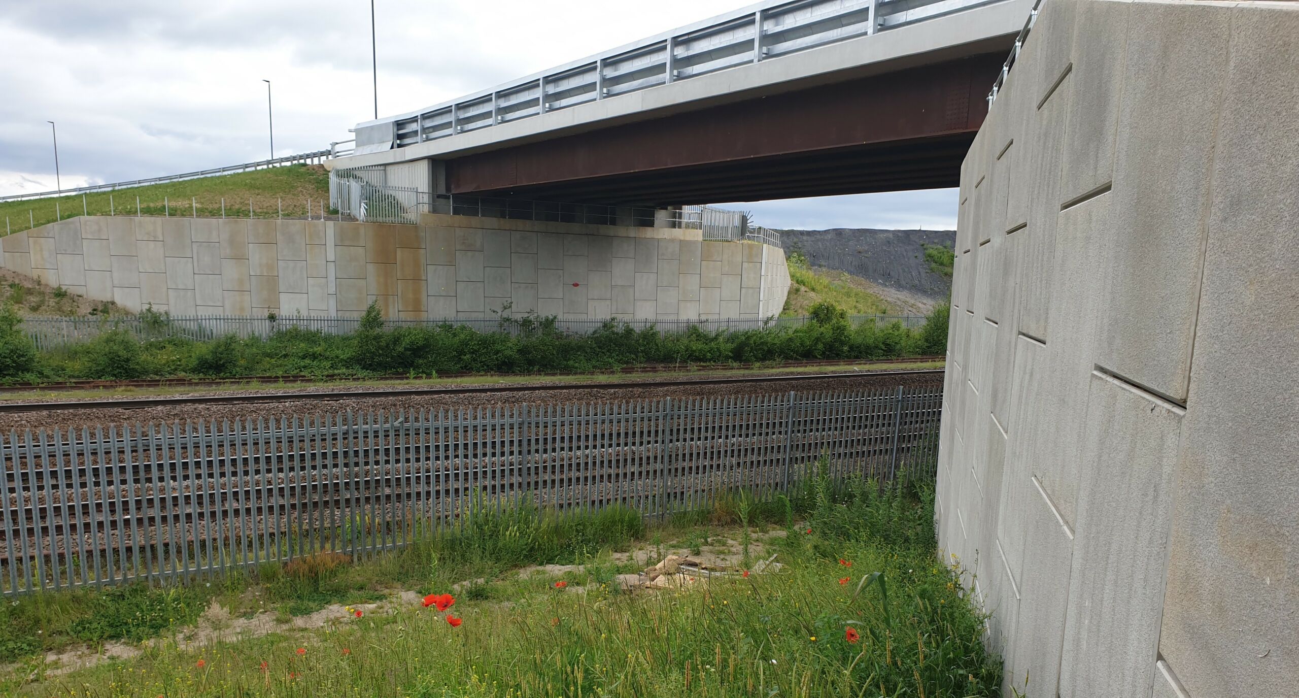 A construction view of the A6 Manchester Airport Relief Road project, showcasing a large retaining wall built using Geoquest Co. Ltd.'s precast concrete facing panels. The image highlights the integration of the road with surrounding infrastructure, illustrating the scale of the project and the innovative design solutions employed for the retaining walls and bridge abutments.